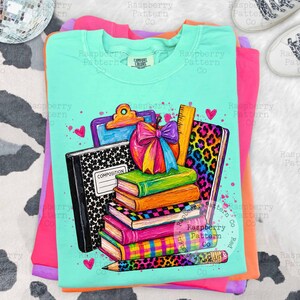 May include: A mint green t-shirt featuring a graphic print of school supplies including a black composition notebook, a stack of colorful books, a clipboard, a ruler, and a pencil, all accented with a vibrant rainbow-colored bow and hearts.