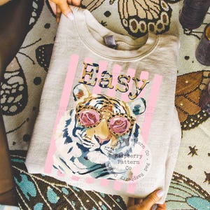 May include: A light gray sweatshirt featuring a tiger wearing pink sunglasses. The word "Easy" is printed above the tiger in a leopard print font, with pink vertical stripes behind the text and tiger.