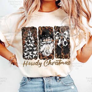 May include: Cream-colored t-shirt featuring a festive design with a Christmas tree, Santa Claus, and a bow, all framed with a leopard print border. The text "Howdy Christmas" is displayed in a gold font.