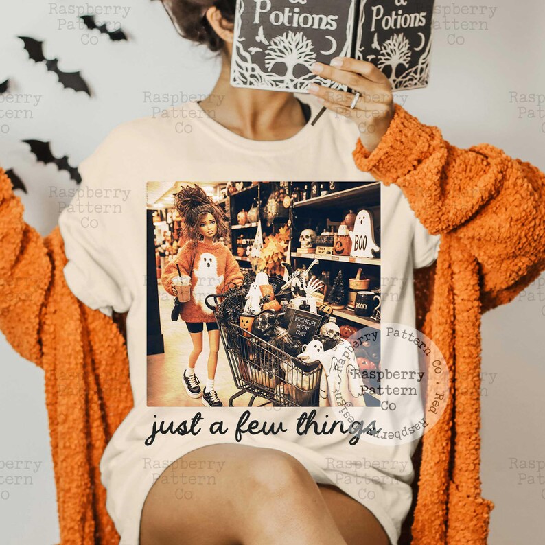 May include: A cream-colored t-shirt featuring a graphic print of a doll in an orange sweater pushing a shopping cart filled with Halloween decorations and the text "just a few things" printed in black script lettering below the image.