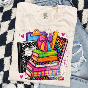 May include: A cream-colored t-shirt featuring a graphic print of a stack of colorful books, a composition notebook, a clipboard, a ruler, and a pencil. The design includes a bright rainbow-colored apple with a bow and leopard print accents.