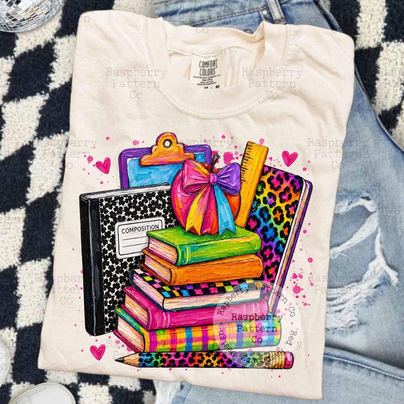 May include: A cream-colored t-shirt featuring a graphic print of a stack of colorful books, a composition notebook, a clipboard, a ruler, and a pencil. The design includes a bright rainbow-colored apple with a bow and leopard print accents.