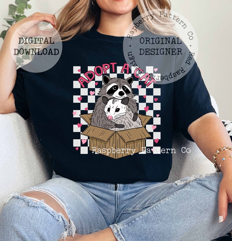 Op de afbeelding: Een zwart T-shirt met een wit-zwarte geruite achtergrond met roze hartjes. De tekst "Adopt a Cat" is geschreven in witte letters met een zwarte omtrek. Een wasbeer en een opossum zitten in een kartonnen doos met de tekst "Raspberry Pattern Co." onderaan.