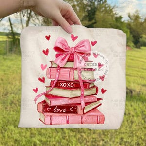 May include: A stack of pink and red books with a large pink bow and ribbon. The books have the words "XOXO" and "love" on them. Red hearts are scattered around the books. The design is on a cream-colored background.
