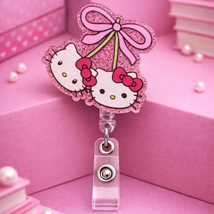 May include: A pink glitter badge reel featuring two Hello Kitty faces with red bows, connected by a green stem and topped with a pink bow. The reel has a clear plastic clip and a retractable cord. The background is a monochromatic pink.