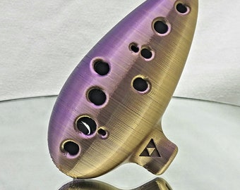 May include: A purple and gold ocarina with multiple finger holes and a mouthpiece. The ocarina has a triangular symbol on the bottom. The instrument is reflected on a mirrored surface.