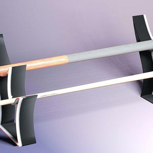 May include: A black and white pool cue rack holding two cues. The rack has a modern design with curved supports. One cue is light-colored with a gray grip, the other is a natural wood color. The rack is designed to hold the cues horizontally.