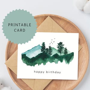 May include: A watercolor birthday card with a mountain and tree design. The card features a green and blue mountain range with evergreen trees and the words "happy birthday". The card is on a wooden tray with a kraft paper envelope.
