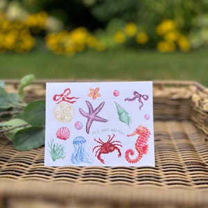 May include: A white card featuring watercolor illustrations of marine life, including a starfish, seahorse, crab, and jellyfish. The card has the text "let's shell-abrate!" in a handwritten font. The illustrations are in shades of red, purple, and blue.