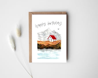 Op de afbeelding: Verjaardagskaart met een aquarel illustratie van een berglandschap, een huis met een rood dak en de tekst "happy birthday". De kaart zit in een bruine envelop, tegen een witte achtergrond met gedroogde bloemen.