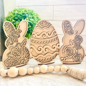 May include: Three wooden Easter decorations: two bunnies and an egg. The bunnies have floral and geometric designs. The egg has a wavy pattern. The decorations are light brown and sit on a white surface with a wooden bead garland.