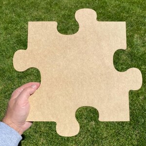 May include: A large, single piece of a puzzle, made of light brown wood. The puzzle piece is held by a hand against a backdrop of green grass. The piece has the classic interlocking shape with four knobs and three indentations.