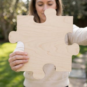 May include: A large, single wooden puzzle piece with a light wood grain finish. The piece is held up by a person, with the background blurred. The puzzle piece has a classic jigsaw shape with four knobs and three corresponding spaces.