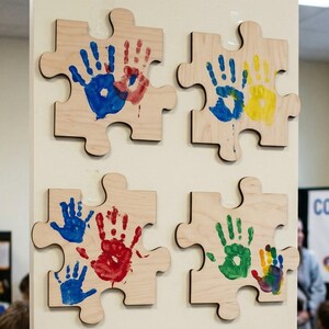May include: Four wooden puzzle pieces with handprints in blue, red, yellow, and green. The puzzle pieces are mounted on a white wall. The handprints are of various sizes and are arranged in different patterns.