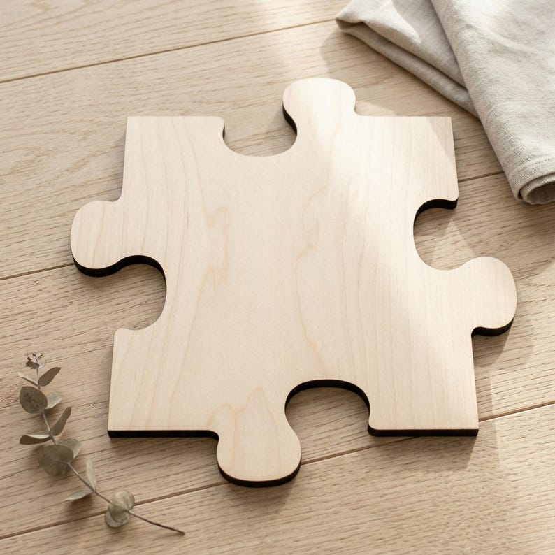 May include: A large, light-colored wooden puzzle piece with a black border. The piece is made of light-colored wood and has a smooth finish. A sprig of greenery and a folded gray cloth are in the background. The puzzle piece is a unique decorative item.