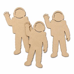 May include: Three flat, light brown, laser-cut wooden astronaut shapes in a standing pose with one arm raised in a wave. Each figure features a rounded helmet outline and a simple, uniform suit design, presented as a set of three distinct cutouts.