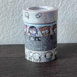 May include: A stack of decorative washi tape rolls. The top roll has a pearlescent finish with small cartoon figures. The middle roll features cartoon characters and text. The bottom roll has a repeating pattern of cartoon figures. The text "THECOFFEEMONSTERCO" is visible.