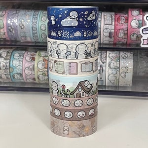 May include: A stack of six washi tapes featuring a repeating pattern of a simple white character with different expressions and activities. The tapes are in a variety of colors, including blue, green, brown, and beige. The tapes are arranged in a stack, with the top tape being blue and the bottom tape being beige.