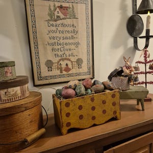 May include: A yellow painted wooden box with red polka dots, filled with colorful Easter eggs. The box is sitting on a wooden table with a framed cross-stitch saying "Dear House, you're really very small--Just big enough for Love--that's all."