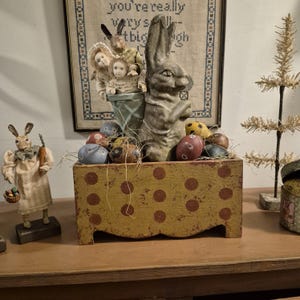 May include: A rectangular wooden box with a distressed mustard yellow finish and dark red polka dots. Inside the box are several painted eggs, a large gray rabbit figurine, and two small dolls. A separate standing rabbit figurine holds a carrot.