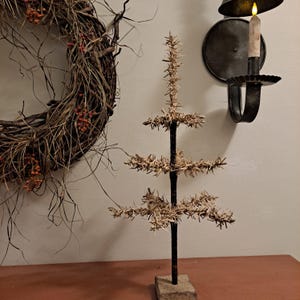 May include: A rustic, handmade Christmas tree with a dark brown trunk and straw-like branches. The tree is mounted on a square, beige base. A wreath with red berries and a candle holder are in the background, creating a warm, seasonal display.