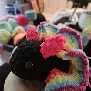 May include: A handmade black crochet creature with rainbow-colored wings and accents. The wings feature shades of pink, blue, yellow, and green. The creature has a button eye and is made of soft yarn.