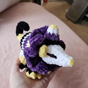 May include: A handmade crochet possum plushie in purple, black, white, and yellow. The plushie has a fluffy texture and is held in a hand. The possum has a small hat and a long tail.