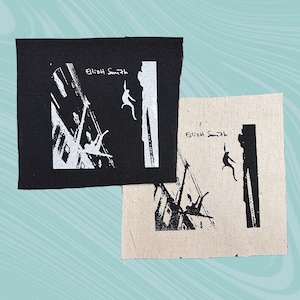 Puede incluir: Dos parches cuadrados con una ilustración en blanco y negro. El diseño muestra una figura cayendo de un edificio, con el texto "Elliott Smith". Un parche es negro, el otro beige.