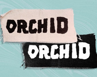 Puede incluir: Dos parches de tela con la palabra "ORCHID" en letras gruesas. Un parche es blanquecino con texto negro, y el otro es negro con texto blanco. Los parches tienen un borde deshilachado y están sobre un fondo azul claro.