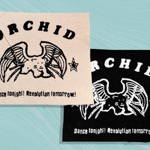 Könnte beinhalten: Zwei Stoffpatches mit dem Wort "ORCHID" in Schwarz auf weißem Hintergrund und Weiß auf schwarzem Hintergrund. Beide Patches zeigen ein stilisiertes Schwarzweißbild einer geflügelten Kreatur mit einem Stern auf jeder Seite. Der Text "Dance tonight! Revolution tomorrow!" ist unter dem Bild auf beiden Patches gedruckt.