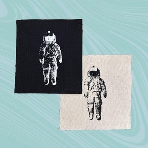 May include: Two square patches featuring a white astronaut design. One patch is black, and the other is a natural beige color. The astronaut is depicted in a standing pose, wearing a spacesuit and helmet. The patches are likely for sewing or ironing onto fabric.