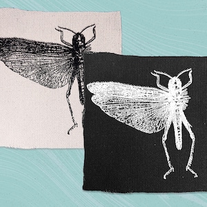 Puede incluir: Dos parches de tela con una imagen en blanco y negro de un saltamontes. Los parches están cosidos juntos, con el saltamontes negro sobre fondo blanco y el saltamontes blanco sobre fondo negro.