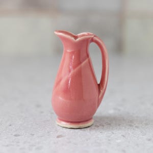May include: A small pink ceramic pitcher with a curved handle. The pitcher has a smooth, glossy finish.