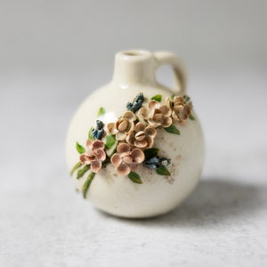 May include: A small white ceramic vase with a handle, decorated with pink and blue flowers. The vase has a rustic, vintage look.