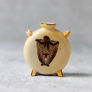 Puede incluir: Un pequeño jarrón redondo de cerámica de color crema con tres patas y dos asas. El jarrón tiene una ilustración marrón de una criatura alada con una estructura esquelética.