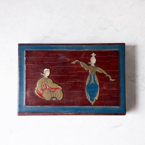 May include: A rectangular wooden box with a dark red interior and a blue border. The box features two stylized figures in gold and blue, one playing a drum and the other dancing. The figures are set against a dark red background.