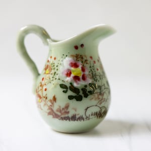 May include: A small, green ceramic pitcher with a floral design. The pitcher has a handle and a spout. The flowers are painted in shades of red, yellow, and white.