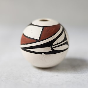 May include: A small, spherical, off-white ceramic bead with a hole at the top. It features a geometric pattern in black and reddish-brown. The design incorporates curved and angular shapes, creating a striking visual.