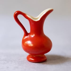 Könnte beinhalten: Ein kleiner, roter Keramikkrug mit einem gebogenen Henkel und einem weißen Rand. Der Krug hat einen bauchigen Körper und steht auf einem kleinen, runden Sockel. Die glänzende Oberfläche reflektiert das Licht und betont die glatten Kurven.