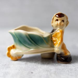 May include: A vintage ceramic planter depicting a boy pushing a wheelbarrow. The wheelbarrow has a leaf design with teal and cream hues. The boy figure is dressed in a yellow jacket and black shoes. A decorative item for the home.