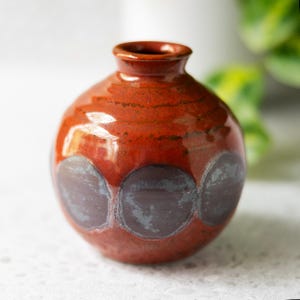 May include: A small, round ceramic vase with a glossy finish. The body is primarily a vibrant burnt orange colour, featuring a series of dark, muted purple circular patterns around the centre. It has a short, narrow neck and a simple, open rim.