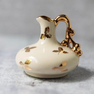 May include: A small, cream-colored ceramic pitcher with a gold handle and rim. The pitcher is decorated with gold leaf clover designs. The handle has a decorative, ornate design. The pitcher is a decorative item.