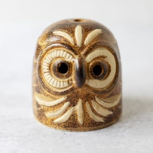 May include: A ceramic owl figurine featuring a speckled brown glaze with cream-coloured carved details. The owl has large, circular eyes with radial lines, a central beak, and stylised feather patterns etched into the surface of the rounded body.