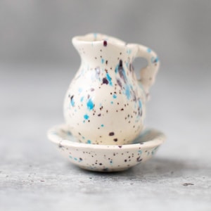 Puede incluir: Una pequeña jarra de cerámica blanca con manchas azules y marrones, sentada sobre un platillo a juego. La jarra tiene un asa y un cuerpo redondeado.