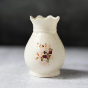 May include: A small, white ceramic vase with a scalloped rim and a floral design featuring butterflies and berries. The vase has a textured, woven-like pattern.