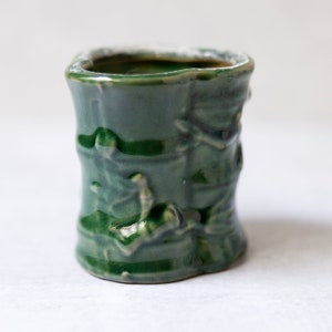 May include: A small, green ceramic pot with a textured, bamboo-like design. The pot is glazed and has a slightly rounded shape.