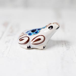 May include: A white ceramic frog figurine with brown and blue painted details. The frog has a hole in its back, suggesting it may be an incense burner.