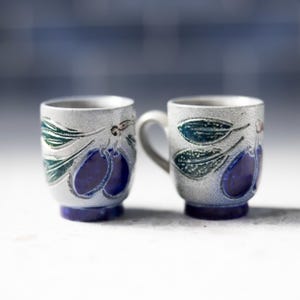 May include: Two ceramic mugs with a light gray textured finish. Each mug features a design of dark blue plums and green leaves. The base of each mug is dark blue. One mug has a handle.
