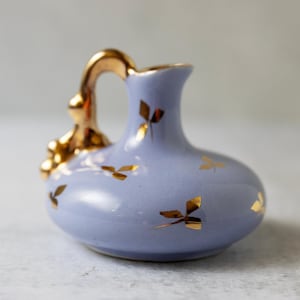 May include: A small, light blue ceramic pitcher with a gold handle and gold leaf accents. The pitcher has a rounded body and a narrow neck. The handle is curved and ornate. The gold leaf accents are scattered across the body of the pitcher.