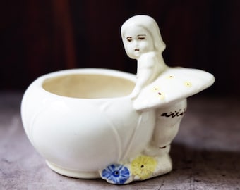 Puede incluir: Maceta de cerámica blanca con forma de niña sosteniendo un hongo. La maceta tiene detalles florales amarillos y azules.